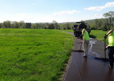 Two workers smooth fresh asphalt on a winding road through a green field, with a truck nearby under clear skies.