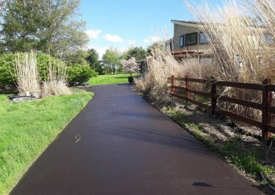 A paved path leads past tall grasses and a wooden fence toward a modern building under a clear blue sky.
