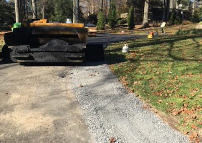 Paving machine on a driveway with gravel, surrounded by trees and fallen leaves.