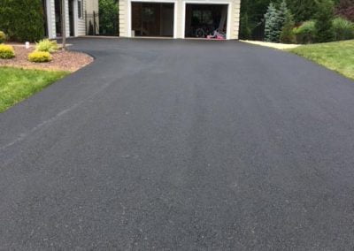 Freshly paved driveway leading to a two-car garage with trees and grass surrounding the area.