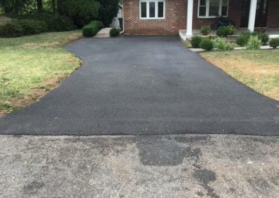 A freshly paved driveway leading to a brick house with a porch, surrounded by grass and trees.