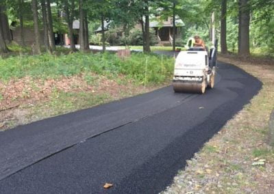Worker operates a roller to lay down new asphalt on a forest road. Trees and houses are in the background.