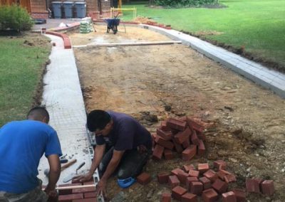 Two people laying red bricks on an outdoor path in front of a house.