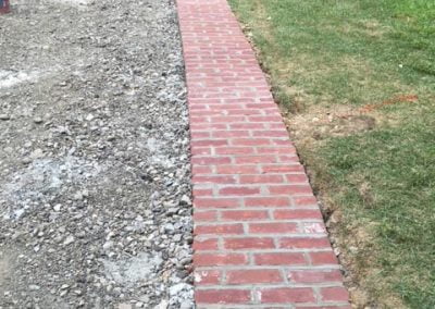 A red brick pathway under construction beside a grassy lawn, with construction tools and a worker in the background.