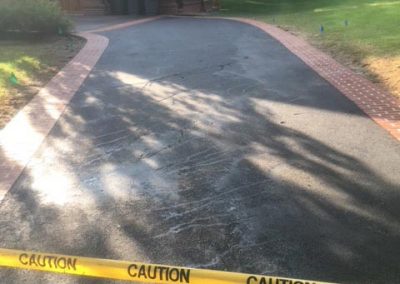 Driveway with caution tape, bordered by bricks, leading to a wooden house surrounded by trees.