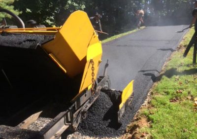 A yellow asphalt paving machine working on a road surrounded by greenery.
