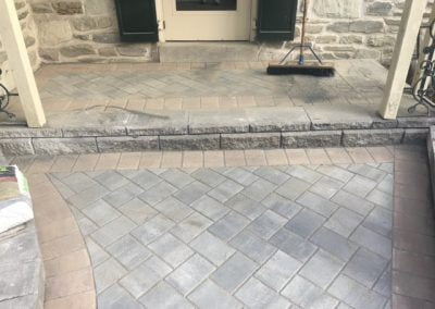 Stone porch with herringbone pattern paving, broom leaning against wall near door.