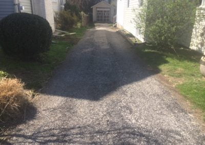 A narrow, sunlit driveway between two houses with shrubs and a small shed at the end.