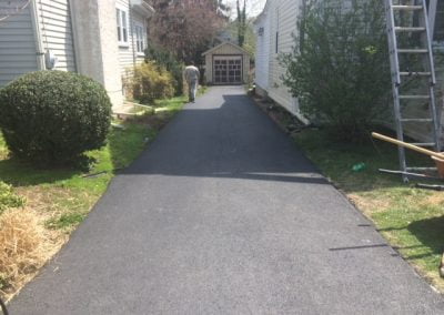 Freshly paved driveway between two houses leading to a small garage; person working in the distance.