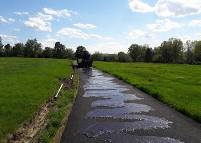 Workers seal a path in a green field under a partly cloudy sky.