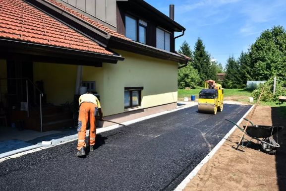 Worker paving a driveway with a roller machine near a yellow house surrounded by greenery.
